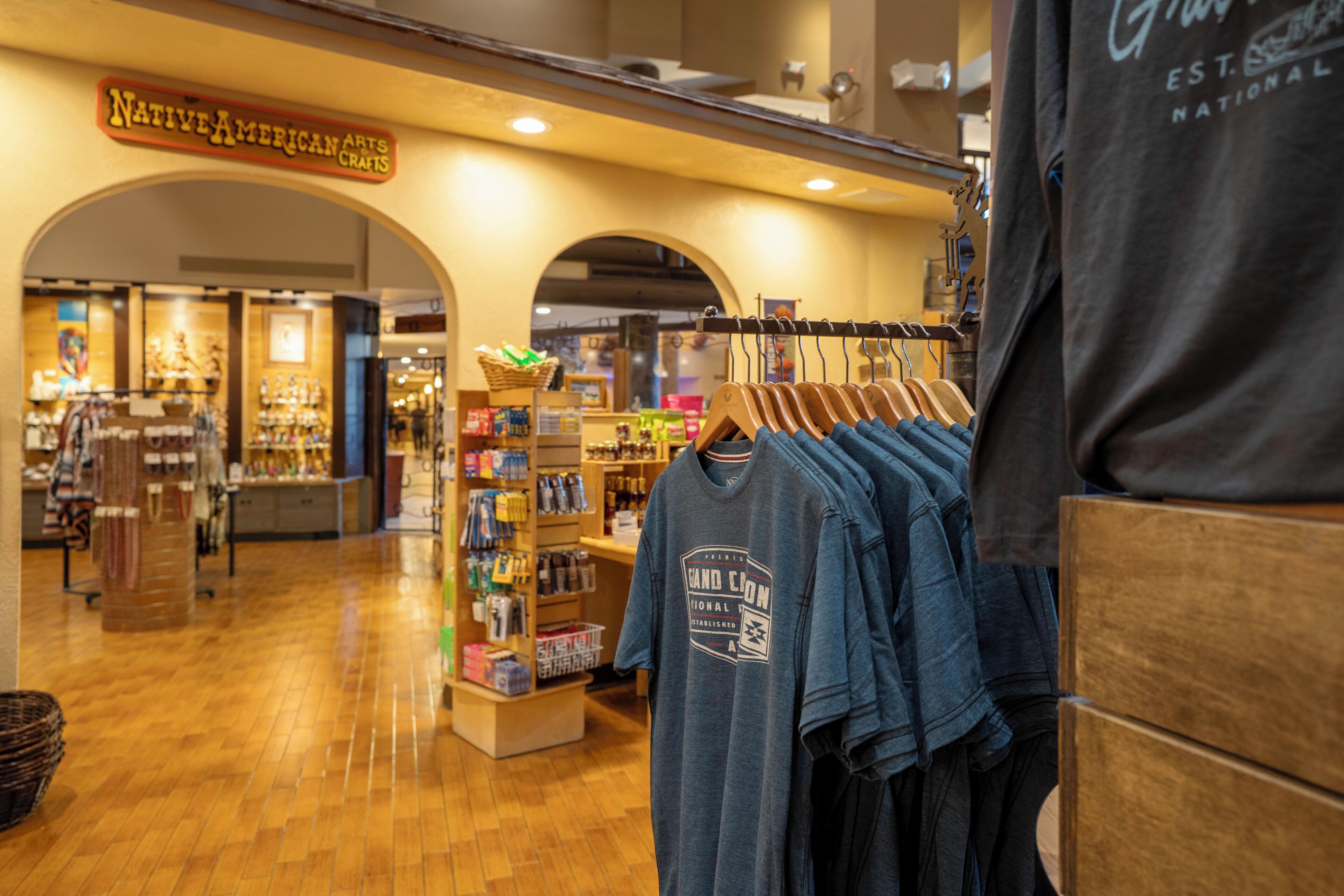 A retail store interior with wooden flooring, a clothing rack with denim shirts, and shelves stocked with various items.