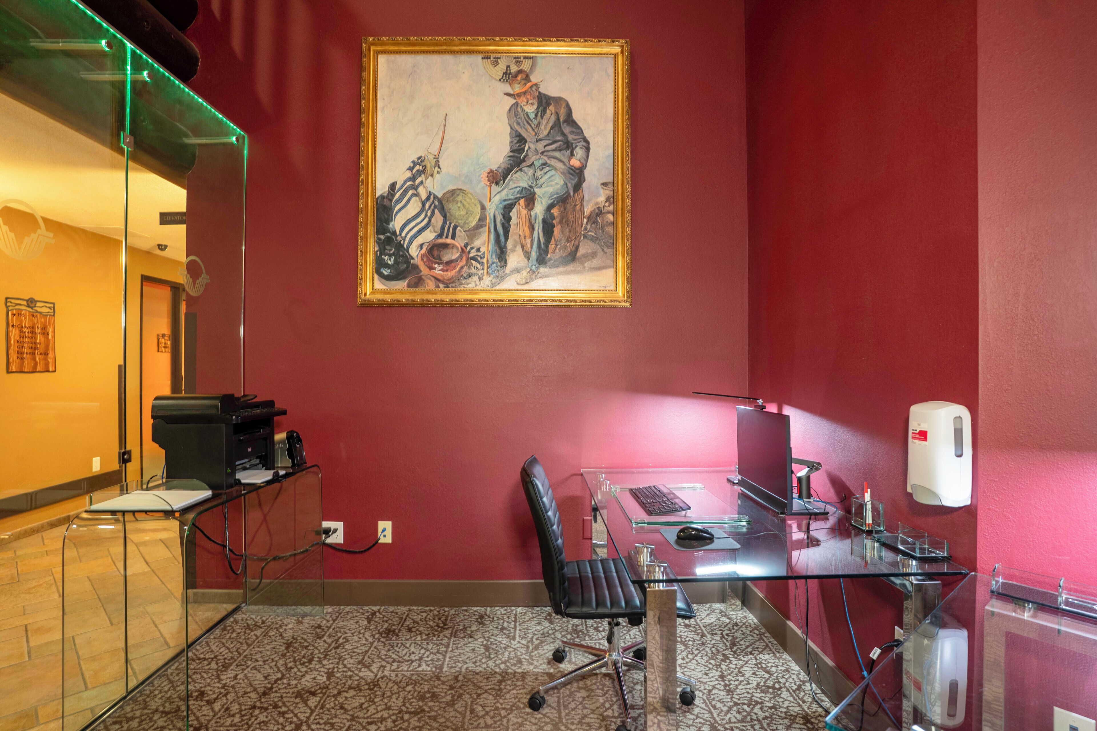 Une salle de conférence avec une table en verre, un bureau avec un ordinateur, une chaise et un tableau accroché au mur.