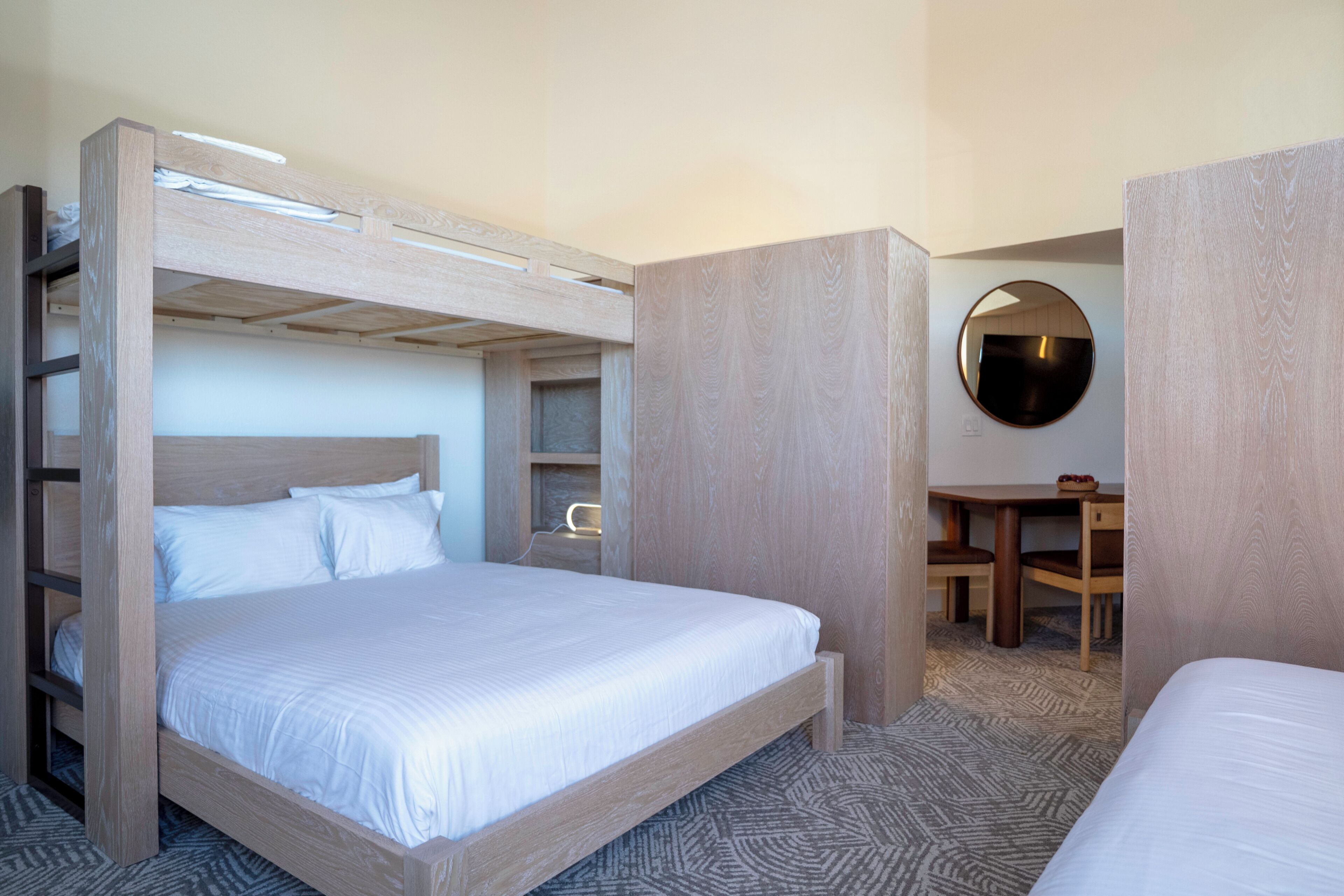 Une chambre d’hôtel avec un lit superposé, un bureau avec deux chaises et un miroir rond accroché au mur.