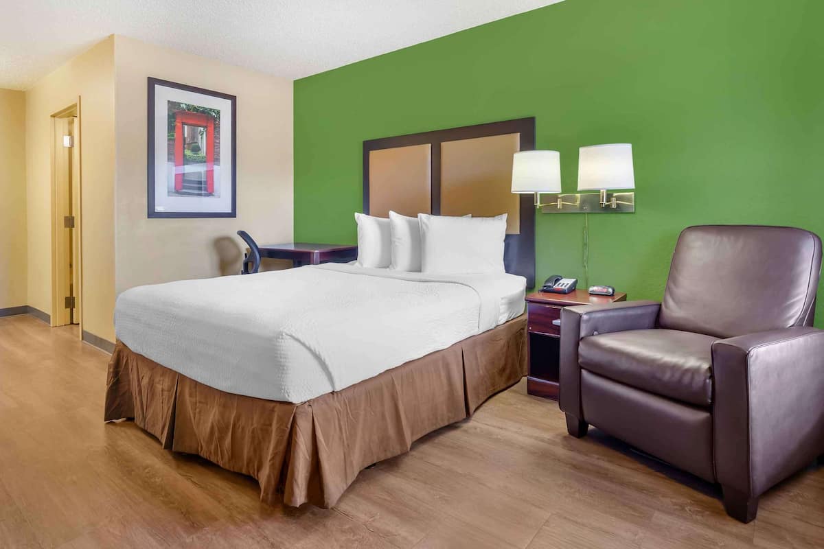 A hotel room with a large bed, a brown leather chair, a desk with a chair, and a green wall.