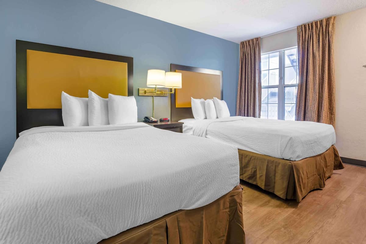 A hotel room with two beds, a nightstand with a lamp, a window with curtains, and a view of the outside.