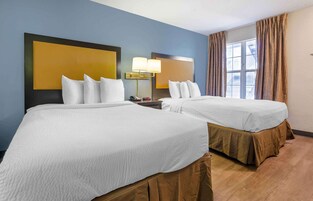 Habitación de hotel con dos camas, una mesita de noche con lámpara, una ventana con cortinas y vistas al exterior.