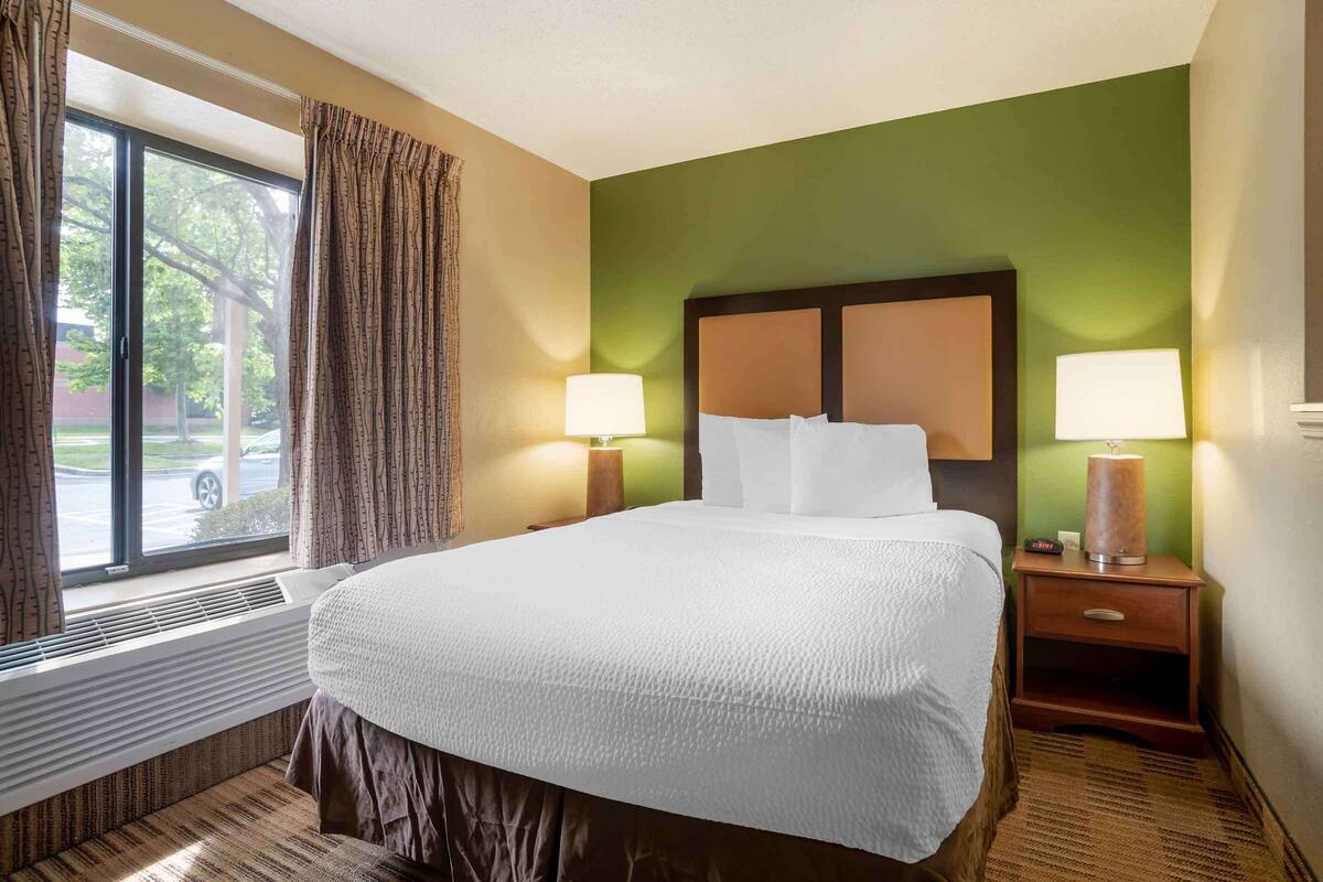 A hotel room with a large bed, two bedside lamps, a window with curtains, and a view of the outside.