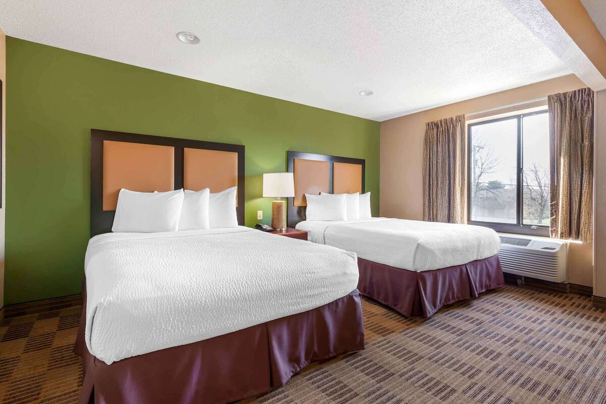 A hotel room with two beds, a large window with curtains, a ceiling light, and a wall-mounted air conditioner.