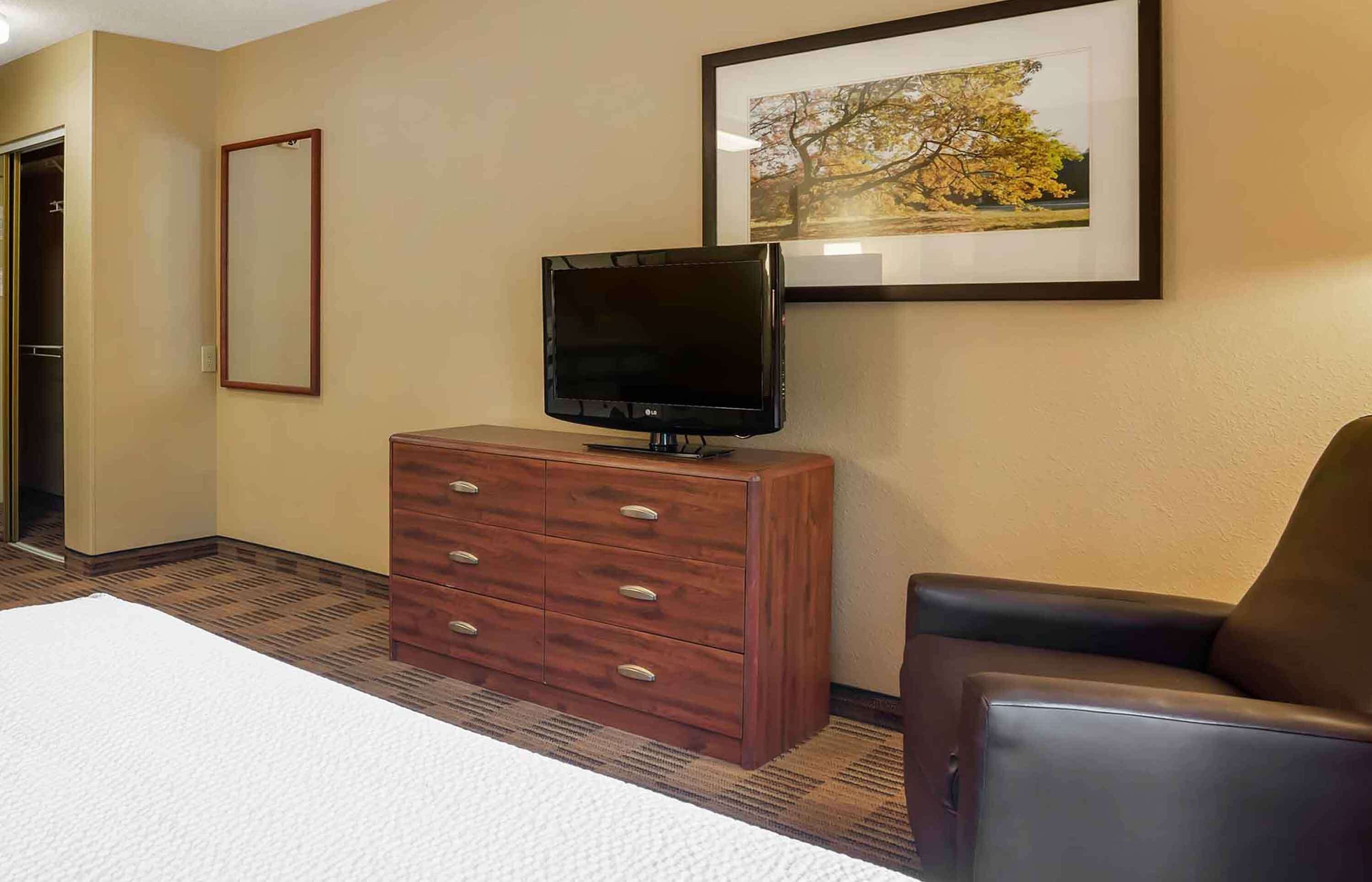 Una habitación de hotel con una cama, un aparador de madera, un televisor, un cuadro en la pared y una silla.