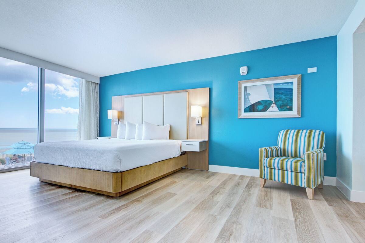A modern hotel room with a large bed, a blue accent wall, a framed picture, and a striped armchair.