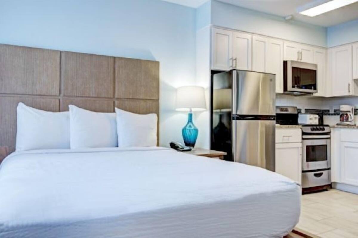 A hotel room with a large bed, a nightstand with a lamp, a refrigerator, a microwave, and a kitchenette with a stove and oven.