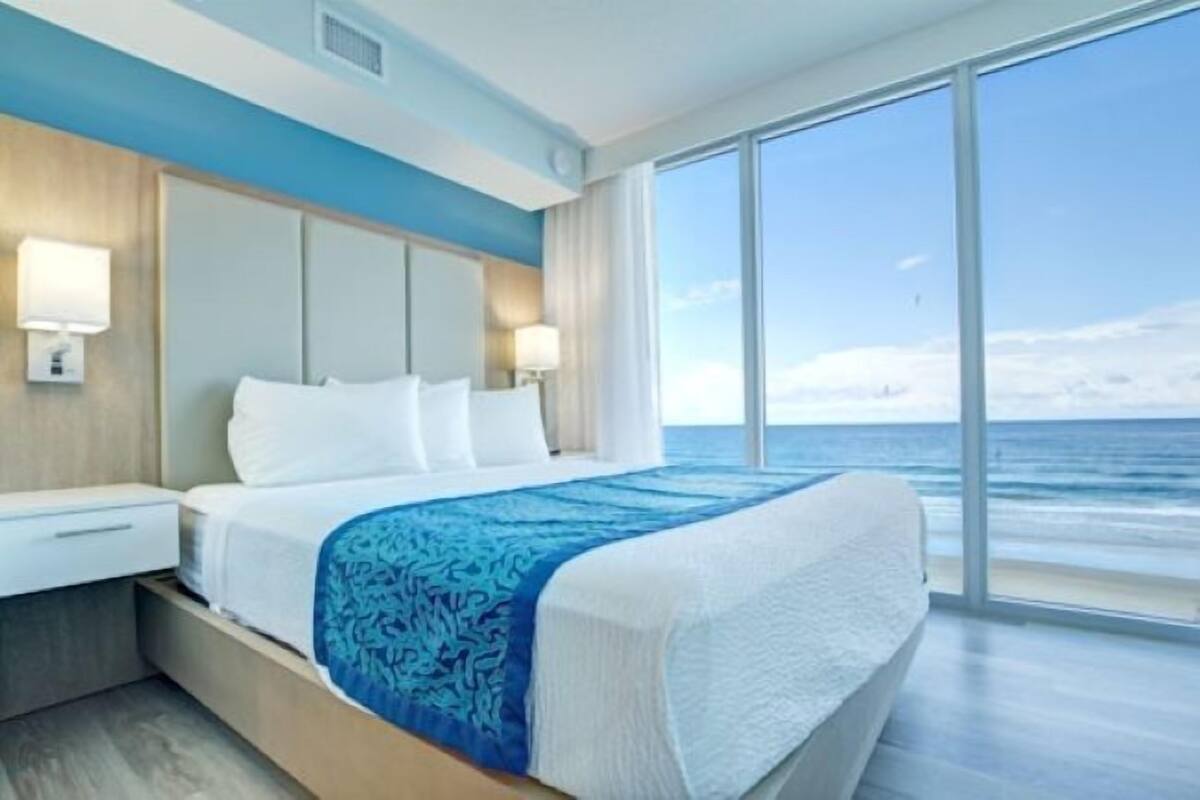 A hotel room with a large bed, a nightstand, a lamp, and a view of the ocean.