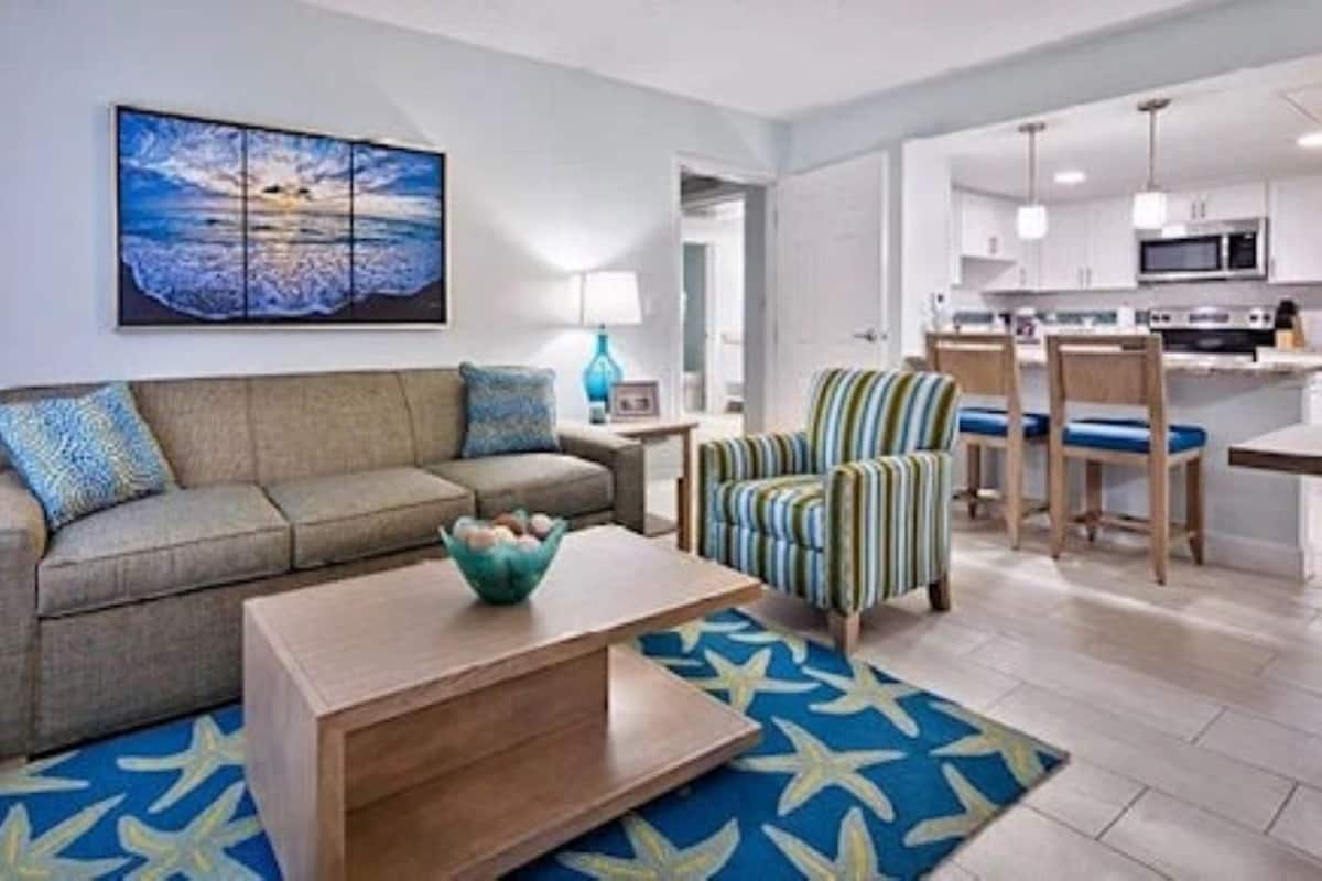 A living room with a sofa, armchair, coffee table, and a painting of a beach scene.