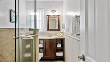 A bathroom with a glass shower enclosure, a wooden vanity with a mirror, and a white tiled floor.