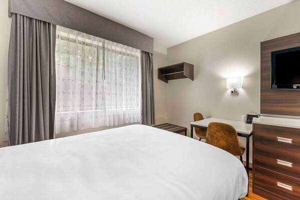 A hotel room with a large bed, a desk with a chair, a TV mounted on the wall, and a small shelf above the desk. - Suburban Studios LaVale-Cumberland (Cumberland)