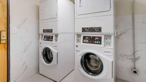 A laundry room with two stacked front-loading washing machines and dryers, marble-tiled walls, and a visible electrical outlet on the wall. - Suburban Studios LaVale-Cumberland (Cumberland)