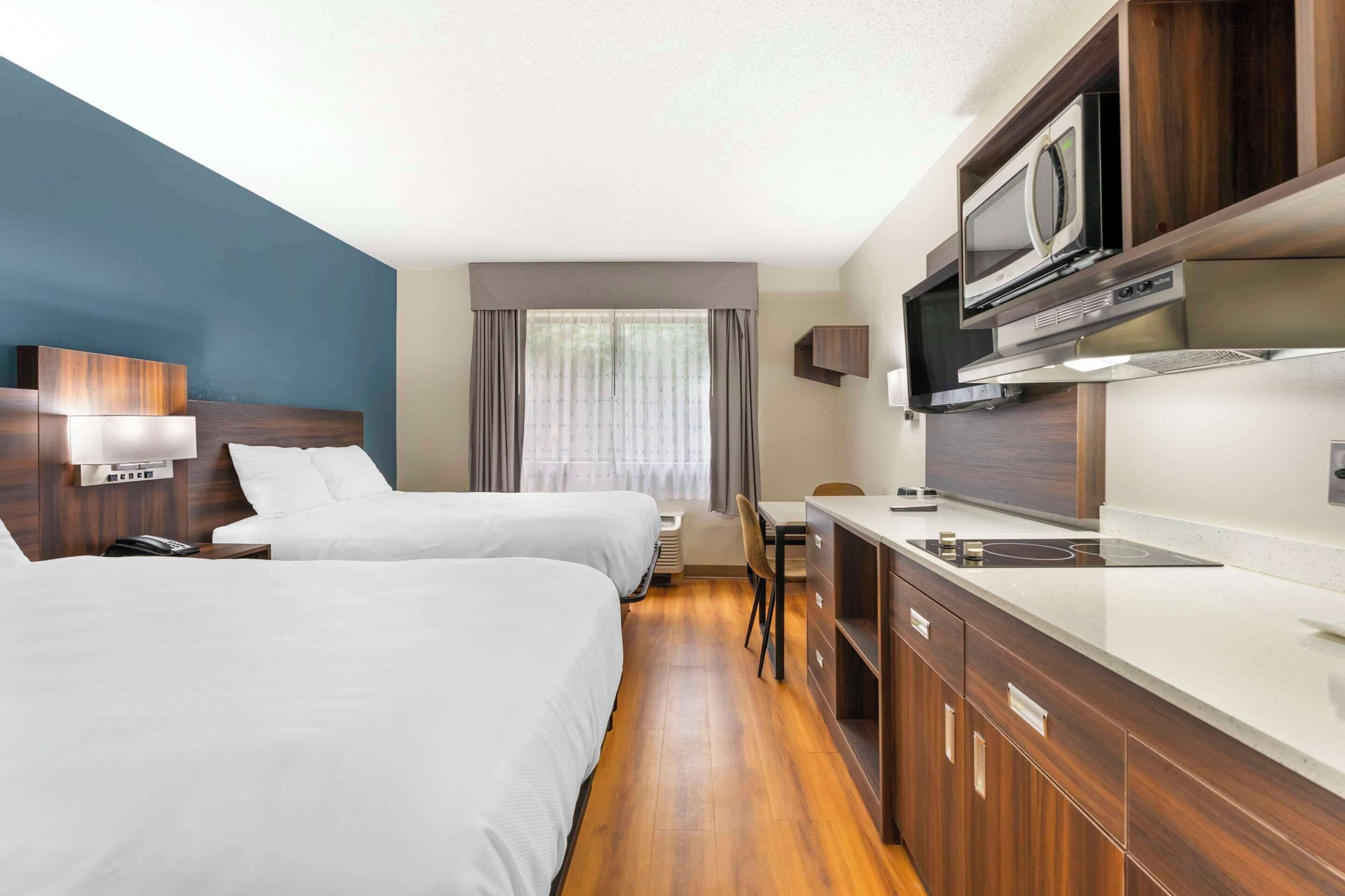 A hotel room with two beds, a kitchenette, a microwave, and a large window with curtains.