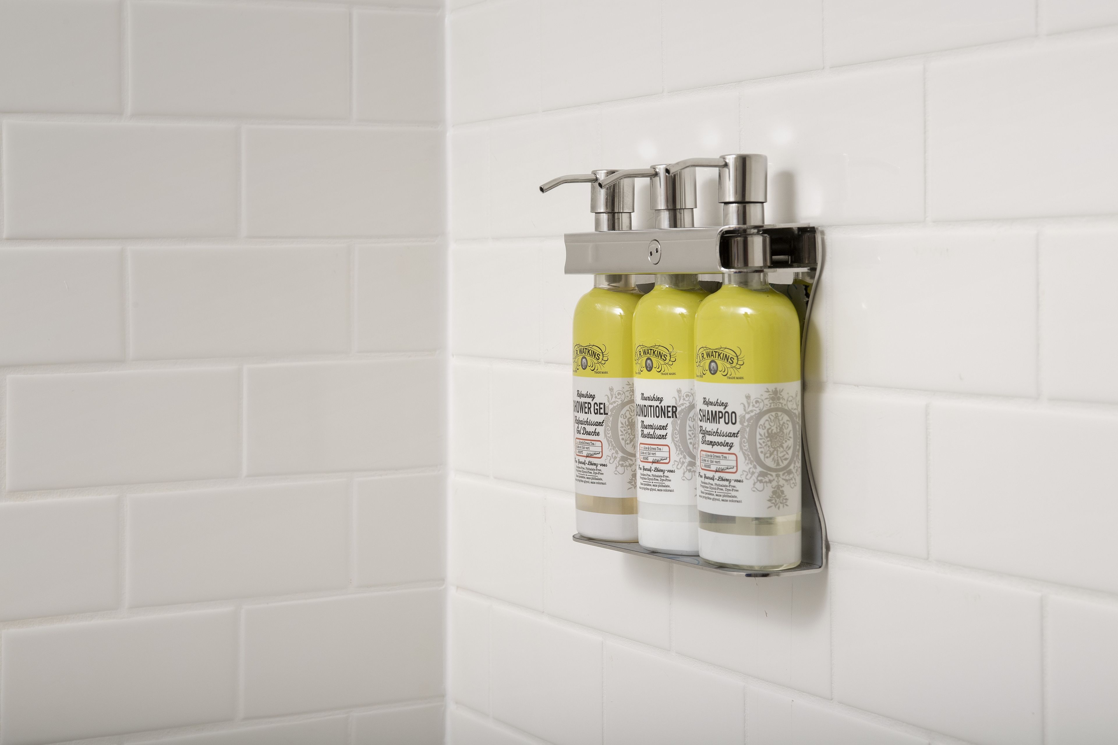 Un baño con paredes de azulejos blancos y un estante con tres botellas de artículos de aseo.