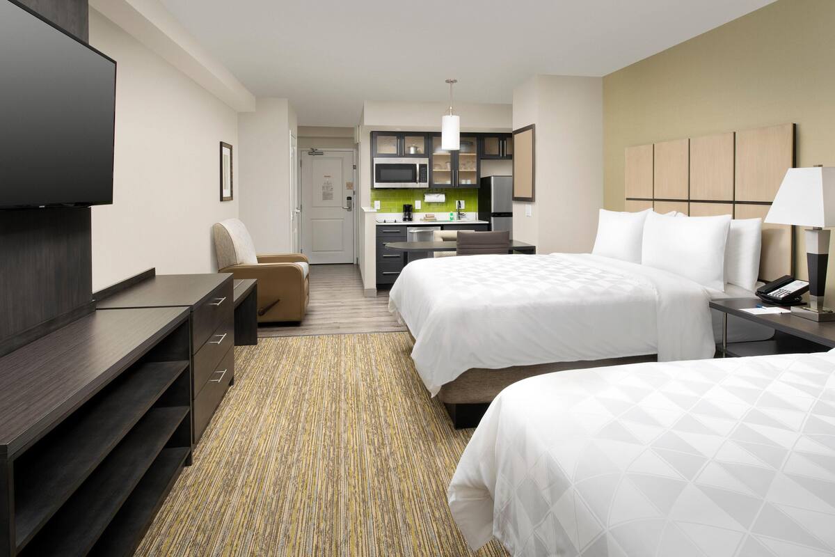 Habitación de hotel con dos camas, televisión, escritorio, silla, kitchenette y baño.