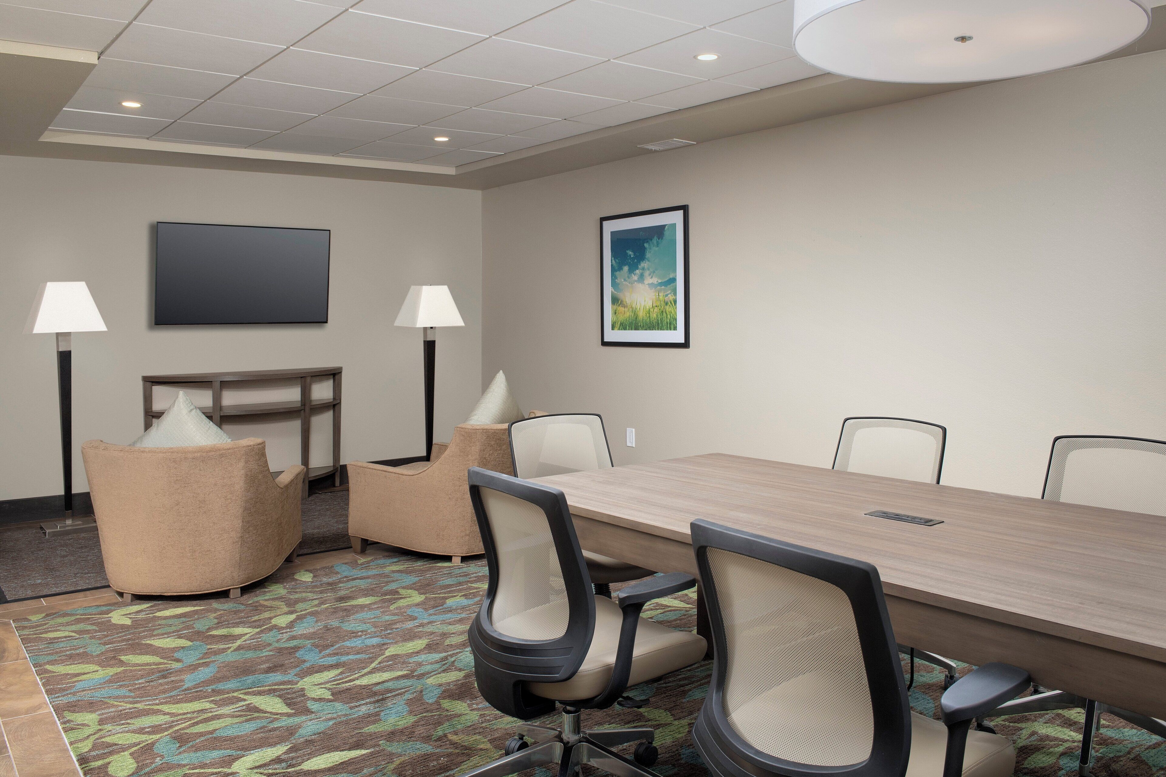 A conference room with a long table, six chairs, two floor lamps, a flat-screen TV, and a framed picture on the wall.