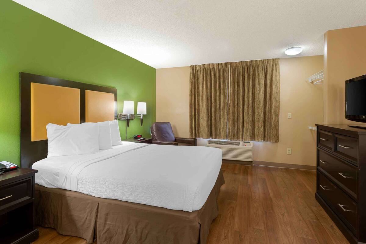 Une chambre d’hôtel avec un lit, un bureau, une chaise, une télévision et une fenêtre avec des rideaux.