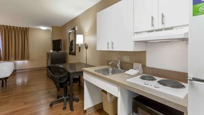 Een kleine hotelkamer met kitchenette, bureau, stoel, televisie en raam met gordijnen.