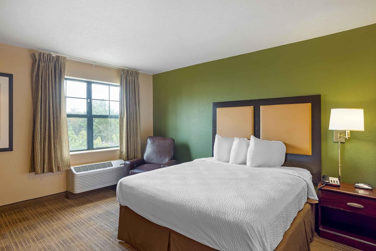 Une chambre d’hôtel avec un grand lit, une chaise, un bureau avec une lampe et une fenêtre avec des rideaux.