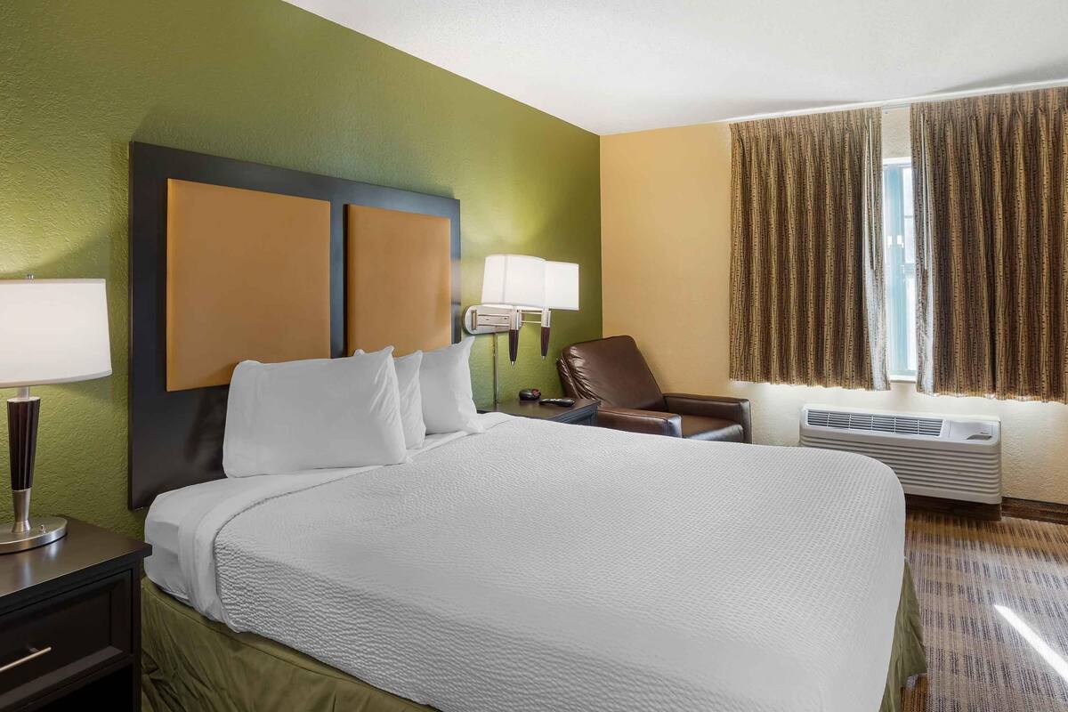 Une chambre d’hôtel avec un grand lit, une table de chevet avec une lampe, une chaise et une fenêtre avec des rideaux.