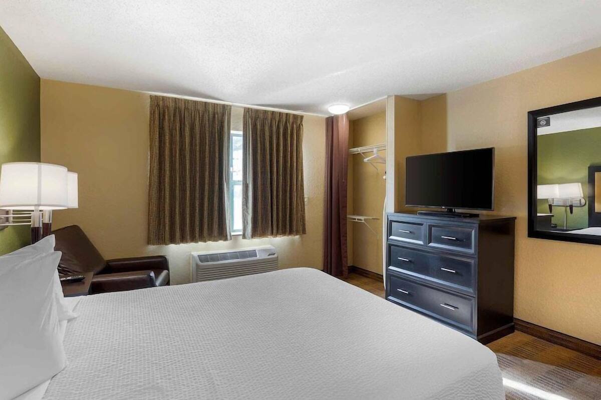 Une chambre d’hôtel avec un grand lit, un téléviseur à écran plat, une commode, une chaise et une fenêtre avec des rideaux.