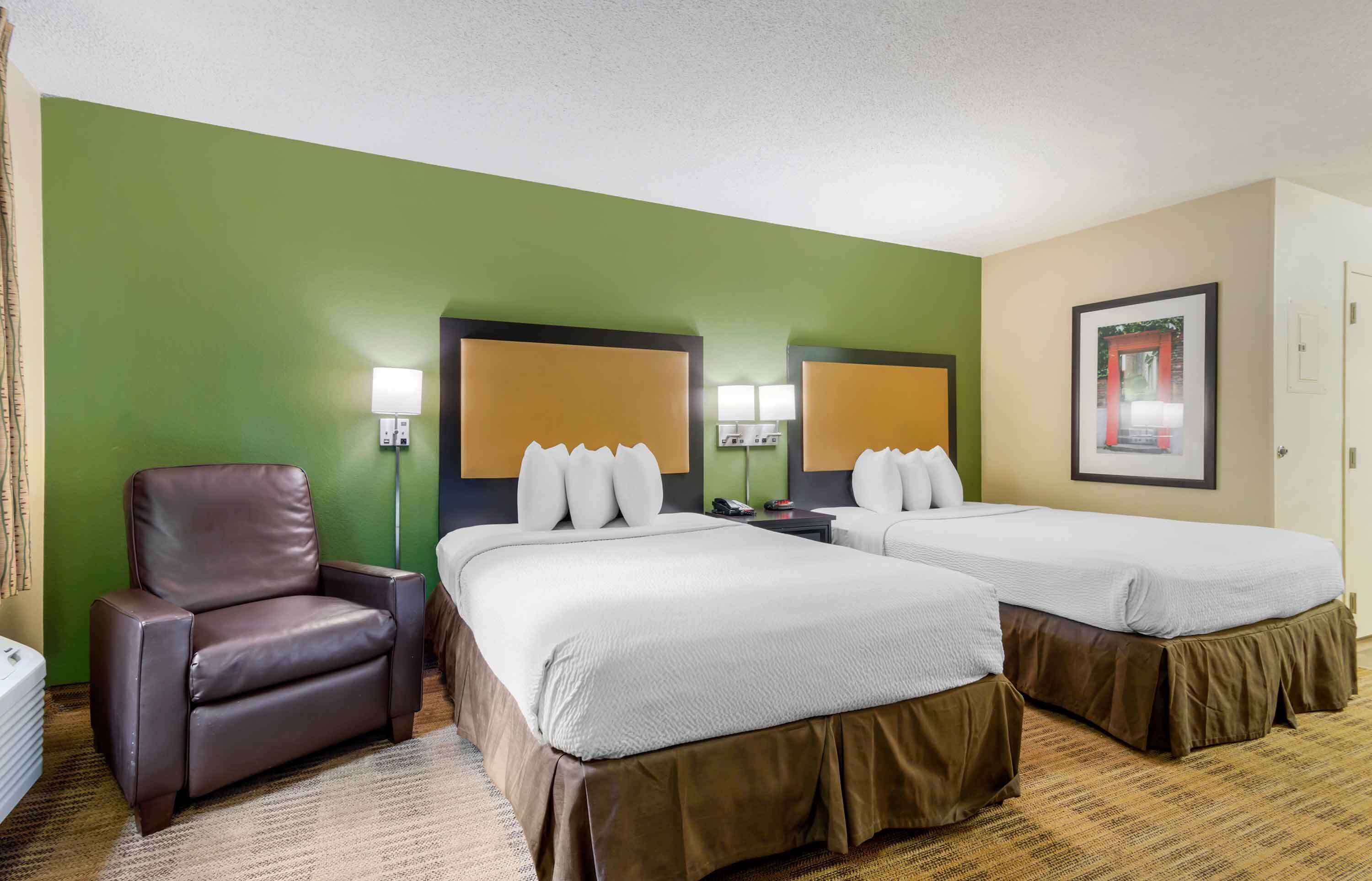 A hotel room with two beds, a brown leather chair, and a green accent wall.