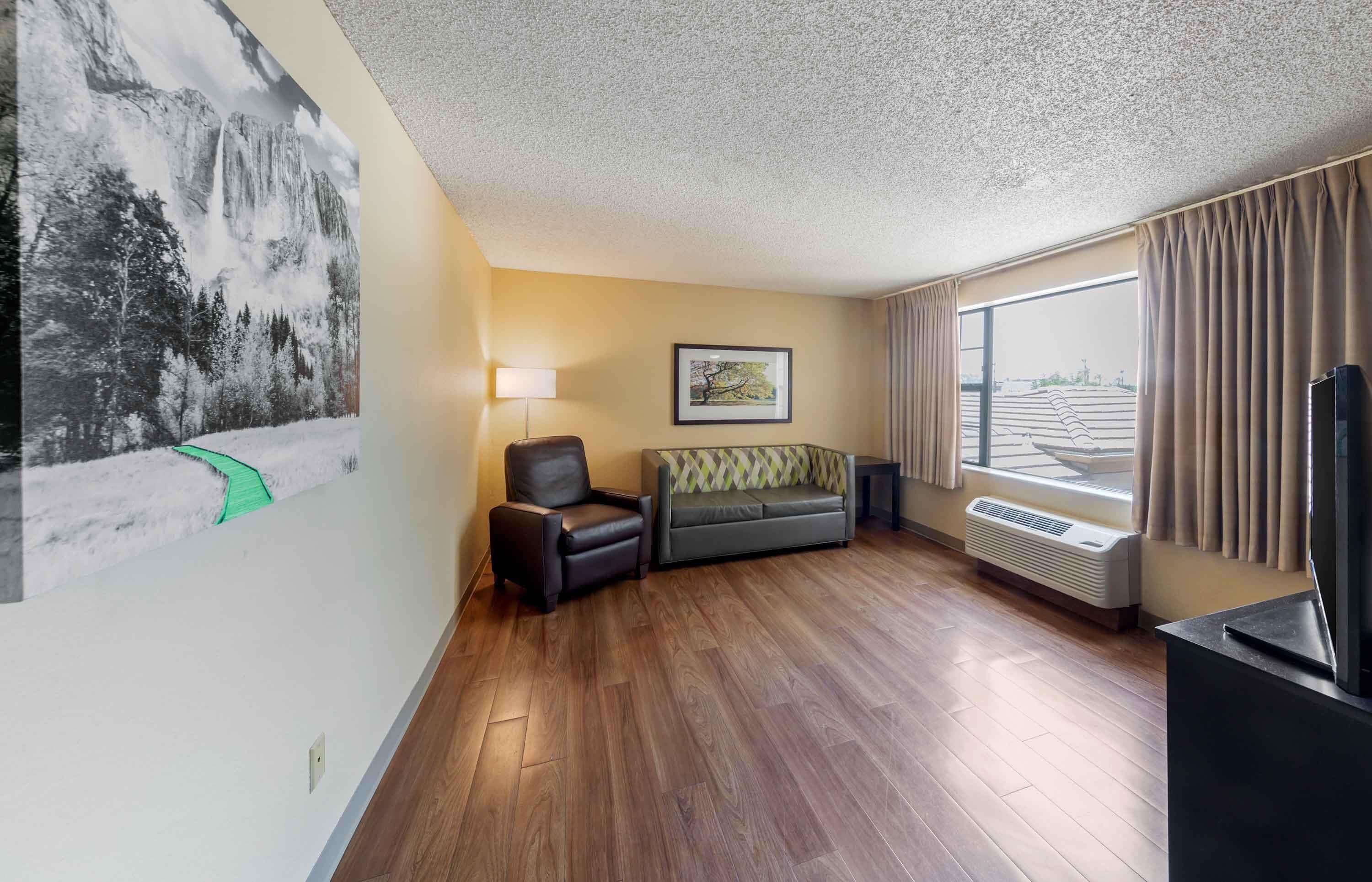A hotel room with a wooden floor, a sofa, a chair, a TV, a window with curtains, and a painting on the wall.