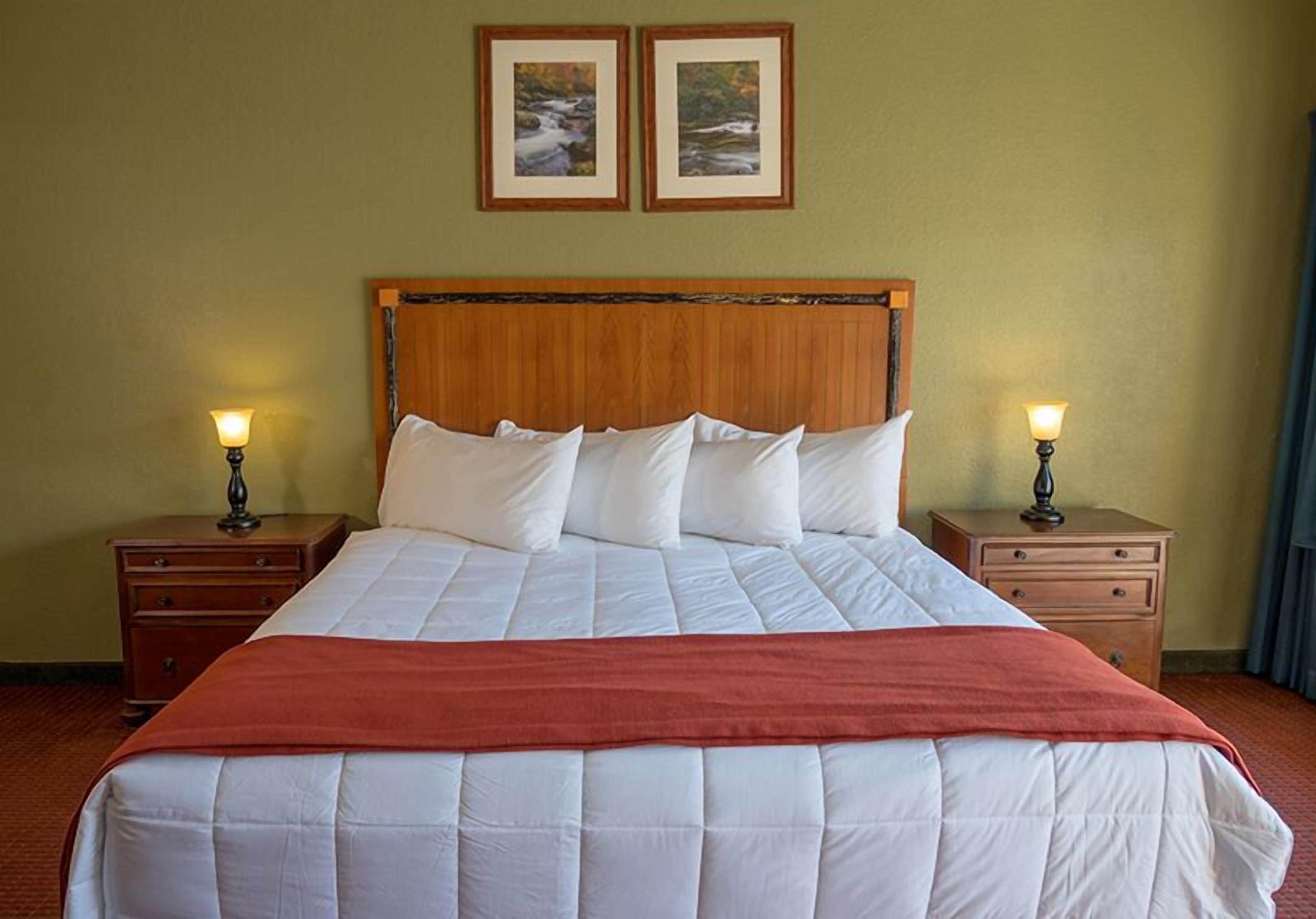 A neatly made bed with white bedding and a red blanket, flanked by two nightstands with lamps, in a room with a wooden headboard and two framed pictures on the wall.