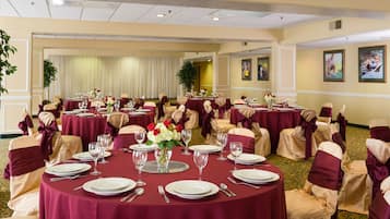A banquet hall with round tables set for dining, each with white plates, silverware, and red flowers.