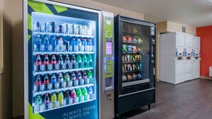 Two beverage vending machines in a room with lockers and a red wall. - Extended Stay America Select Suites - Orlando - Conven Ctr - Sports Complex (Orlando)