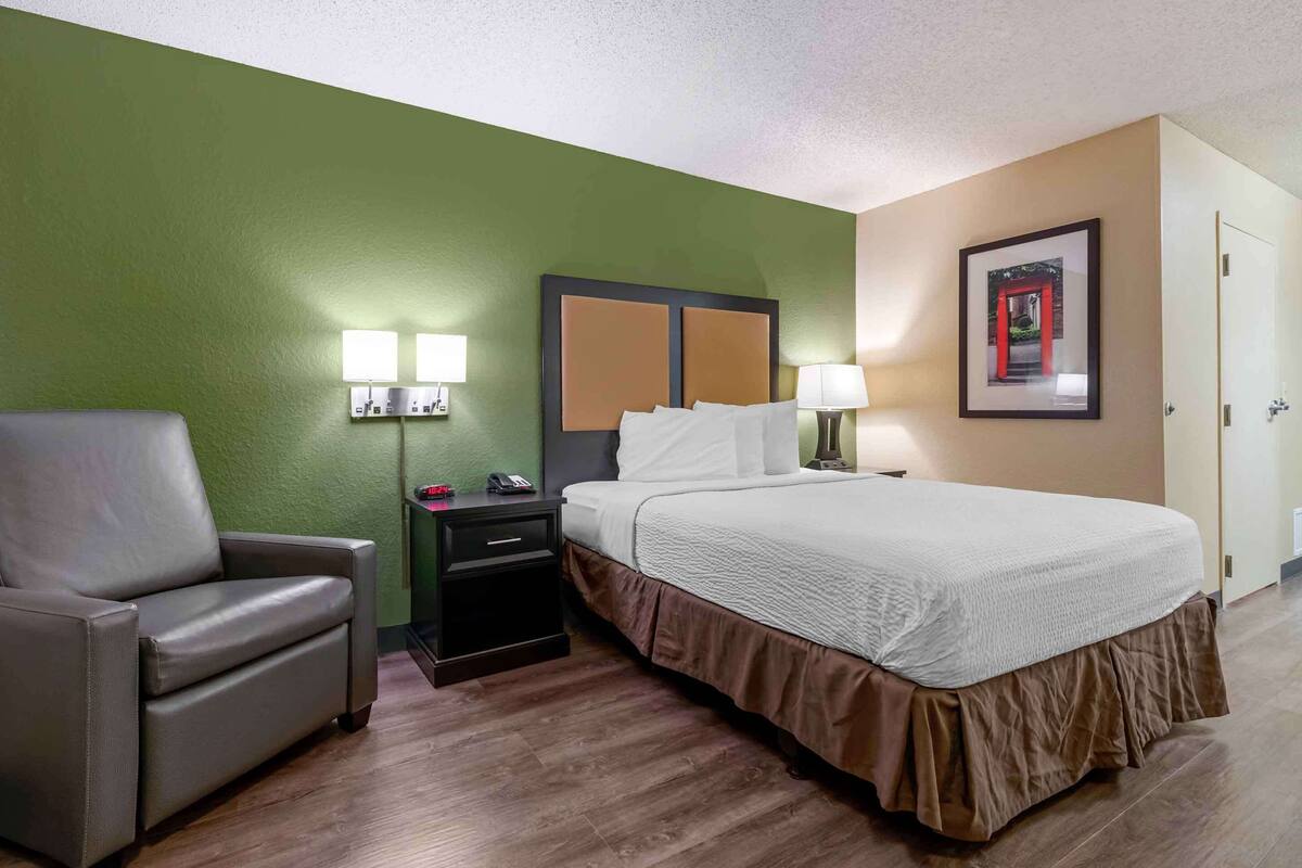 A hotel room with a bed, a chair, a nightstand with lamps, and a framed picture on the wall.