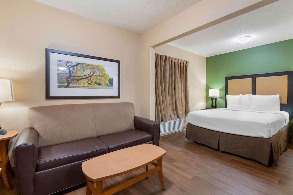Ein Hotelzimmer mit Bett, Sofa, kleinem Tisch, einem gerahmten Bild an der Wand und einem Fenster mit Vorhängen.