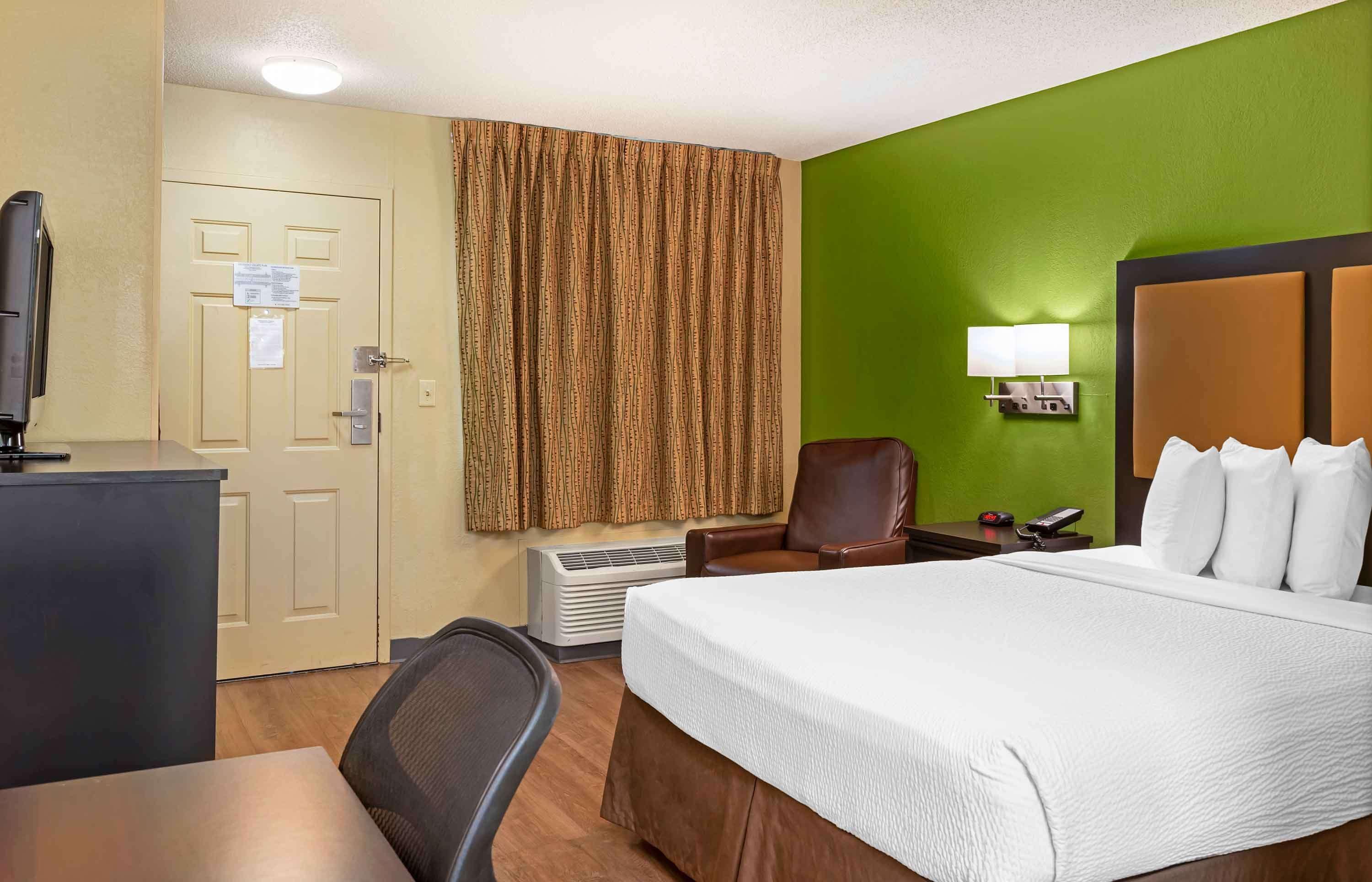 Ein Hotelzimmer mit Bett, Schreibtisch, Stuhl, Fernseher, Lampe und einem Fenster mit Vorhängen.