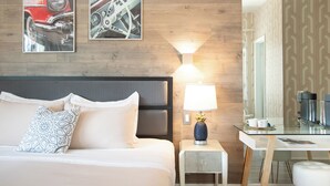 A hotel room with a bed, a nightstand with a lamp, a desk, and two framed pictures on the wall. - Century Hotel (Miami Beach)