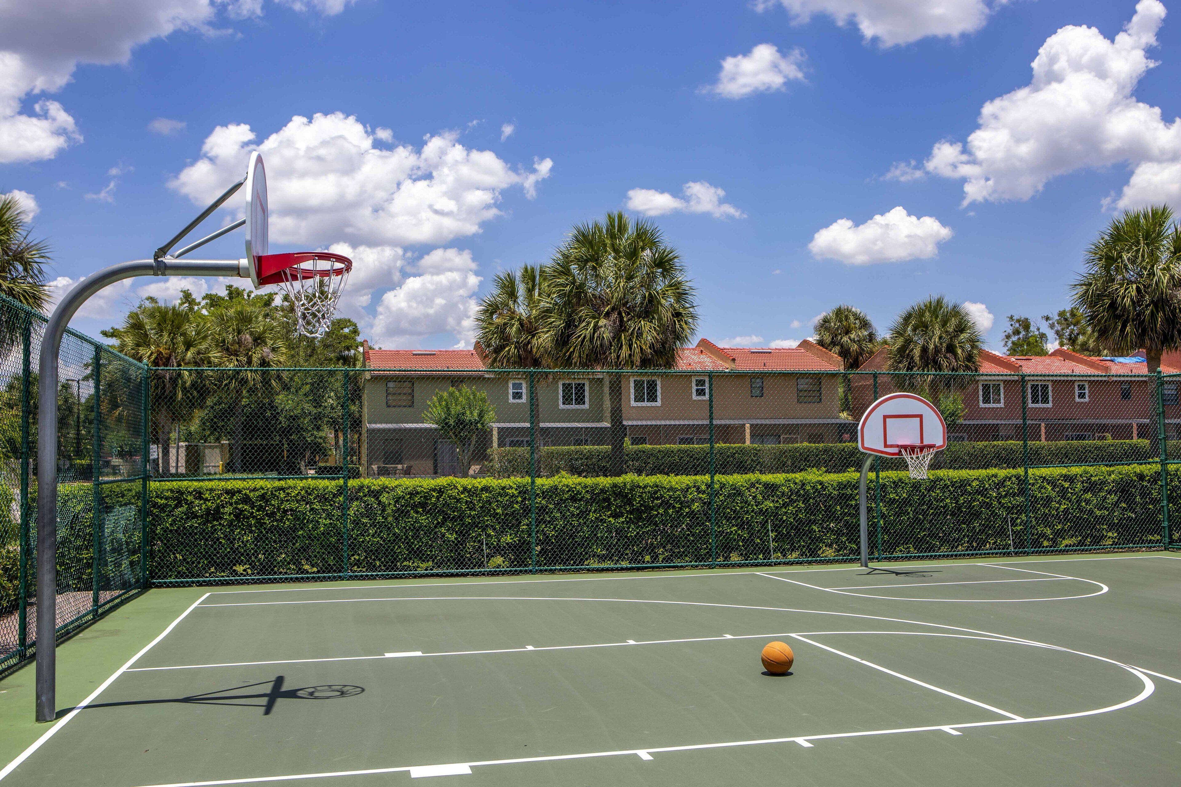An outdoor basketball court with a hoop, a basketball, and a clear blue sky with clouds.