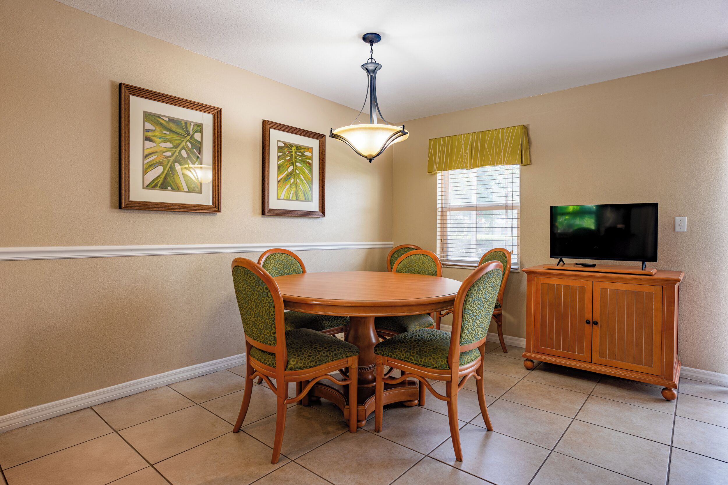 A dining area with a round wooden table, four chairs, a television on a wooden cabinet, and framed artwork on the wall.