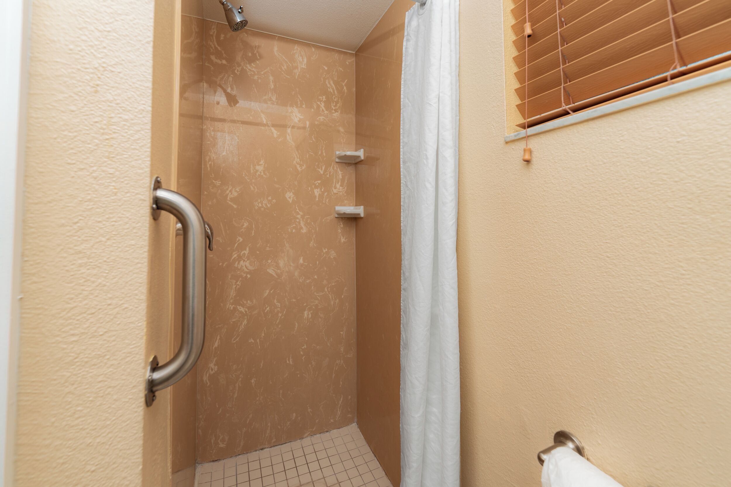 Un baño con ducha, barra de apoyo, ventana con persianas y un perchero para toallas.
