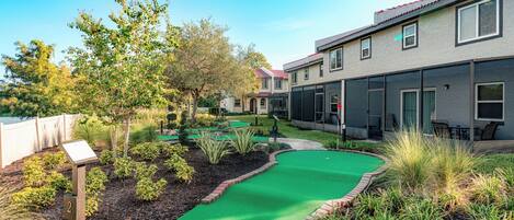 Un barrio residencial con un campo de golf de césped artificial, un sendero de ladrillo y una fila de casas con ventanas amplias y un espacio de patio cubierto.
