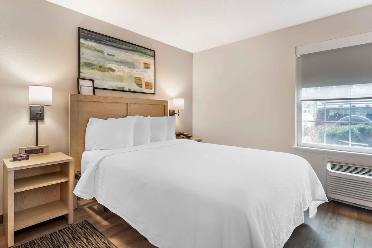 A hotel room with a large bed, a wooden headboard, two bedside tables with lamps, a painting on the wall, and a window with a view of the outside.