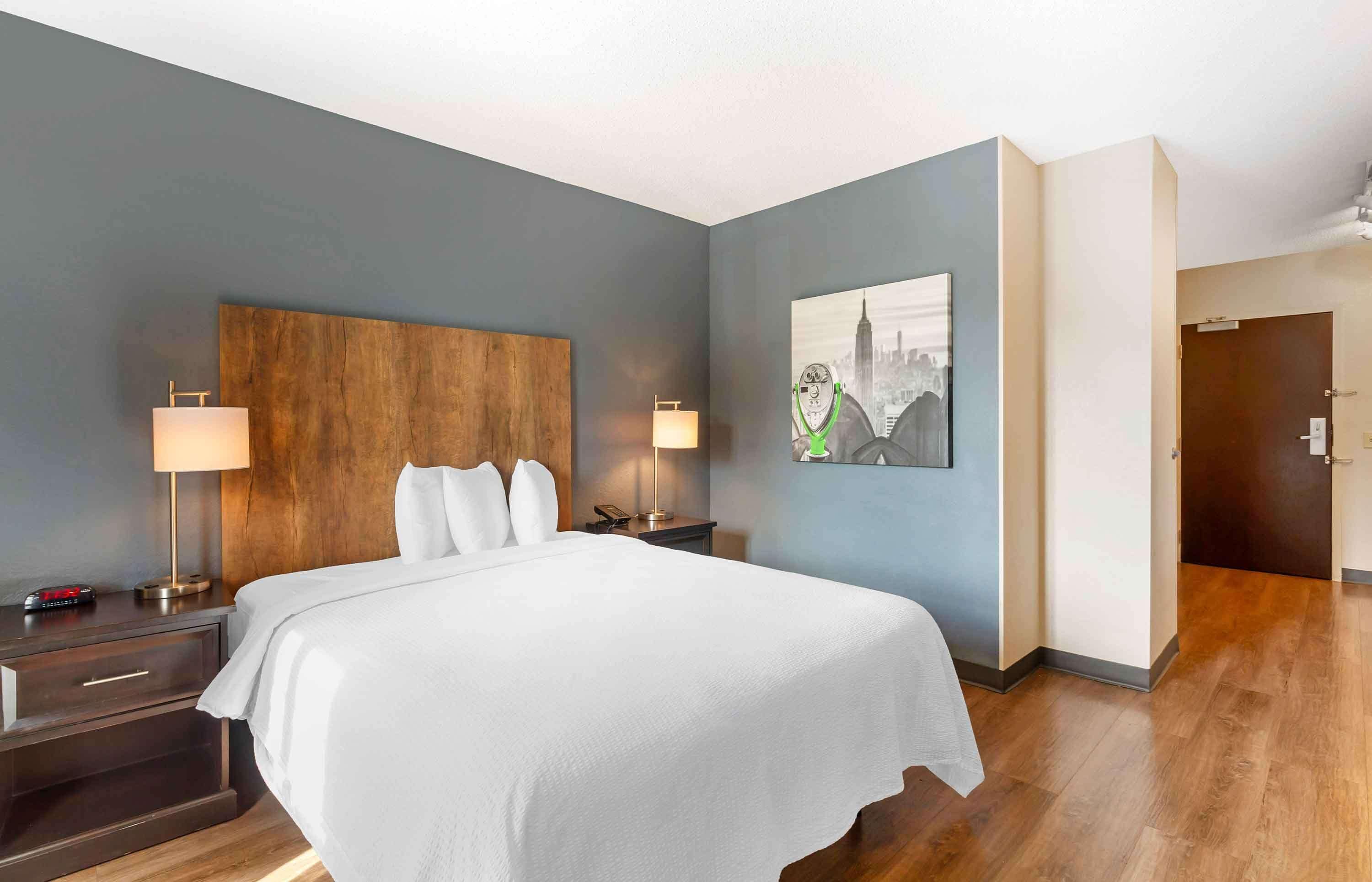 Une chambre d’hôtel avec un grand lit, deux tables de chevet avec des lampes, une tête de lit en bois et un tableau représentant un paysage urbain accroché au mur.