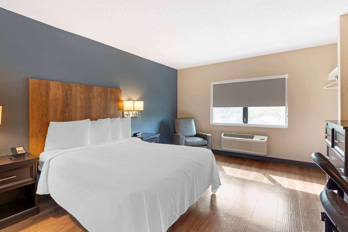 Ein Hotelzimmer mit einem großen Bett, Nachttischlampen, einem Sessel, einem Schreibtisch und einem Fenster mit Jalousien.