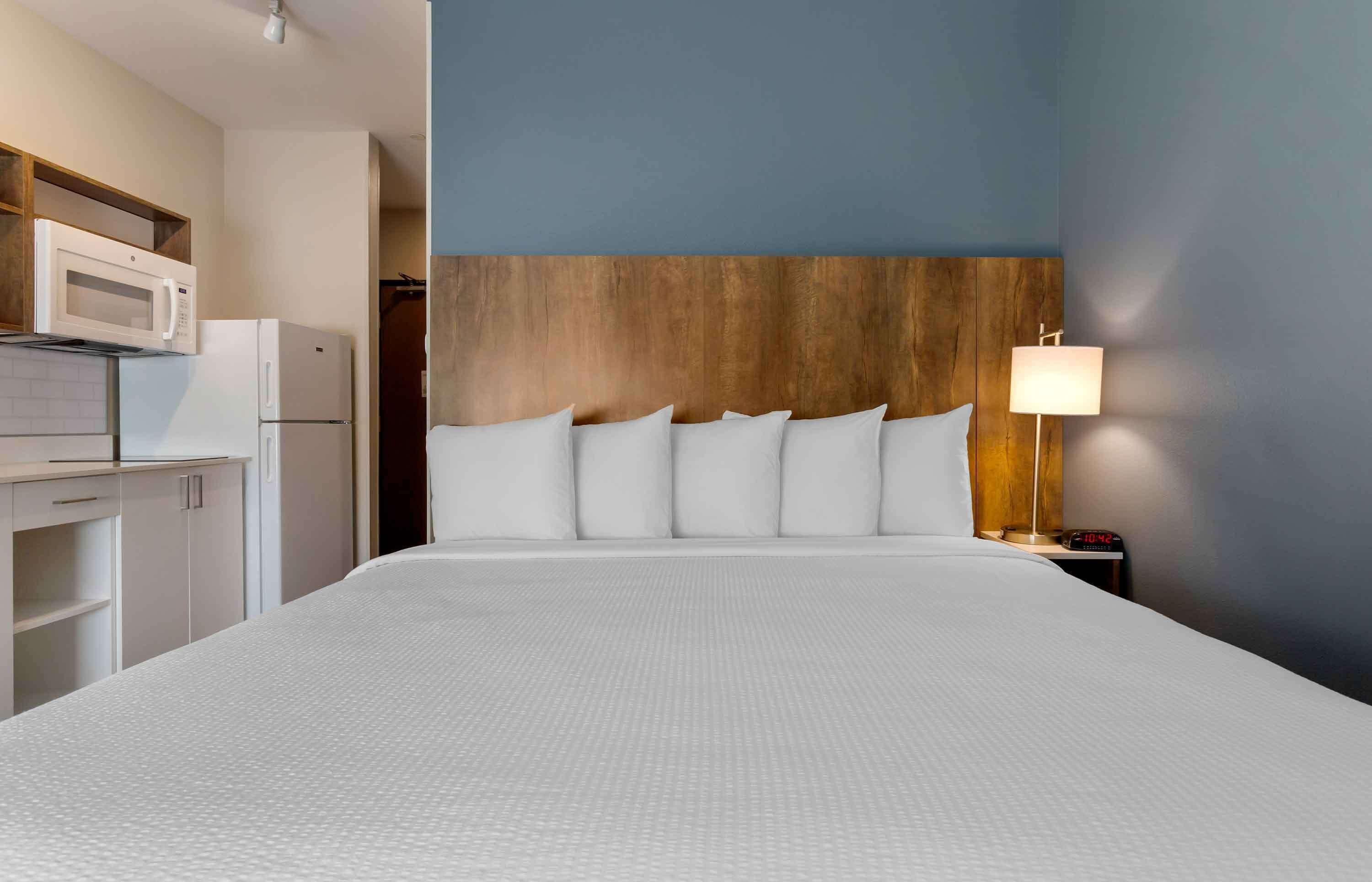 Een hotelkamer met een groot bed, een houten hoofdbord, een nachtkastje met een lamp en een kitchenette met een magnetron en koelkast.