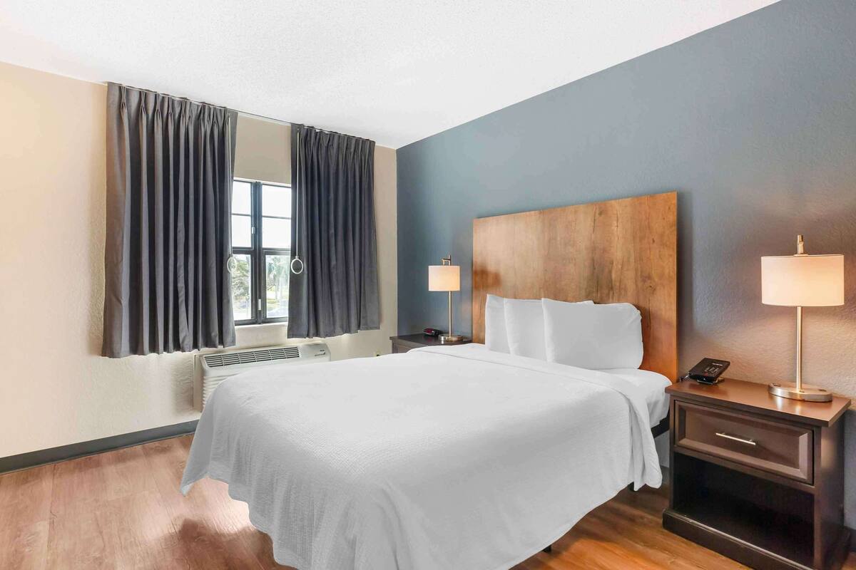 Ein Hotelzimmer mit einem großen Bett, einem hölzernen Kopfteil, zwei Nachttischen mit Lampen, einem großen Spiegel und einem Fenster mit Vorhängen.