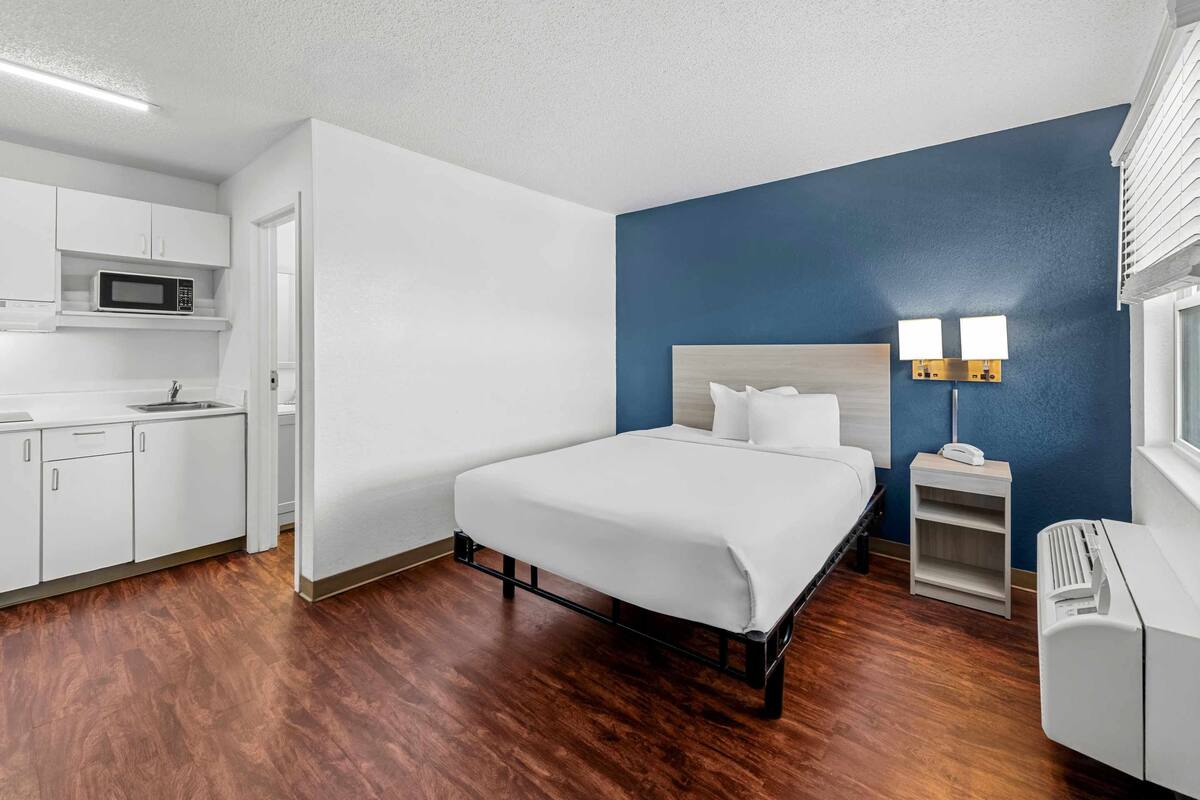 Ein Hotelzimmer mit Bett, einem kleinen Nachttisch mit Lampe, einer Mikrowelle in der Küchenzeile und einem Fenster mit Jalousien.