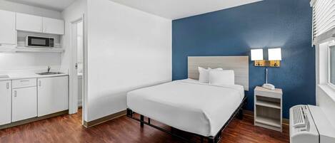 A hotel room with a bed, a small bedside table with a lamp, a microwave in the kitchenette, and a window with blinds.