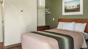 A bedroom with a bed, a nightstand, a wall-mounted light switch, and a framed picture on the wall. - HomeTowne Studios By Red Roof Eugene - Springfield (Springfield)