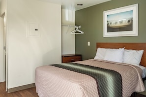 A bedroom with a bed, a nightstand, a wall-mounted light switch, and a framed picture on the wall. - HomeTowne Studios By Red Roof Eugene - Springfield (Springfield)