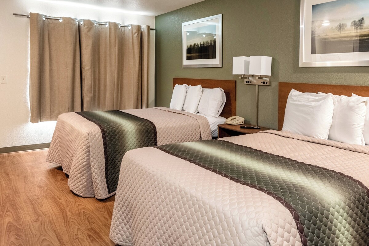 A hotel room with two beds, a wooden floor, a nightstand with a lamp, and a framed picture on the wall.