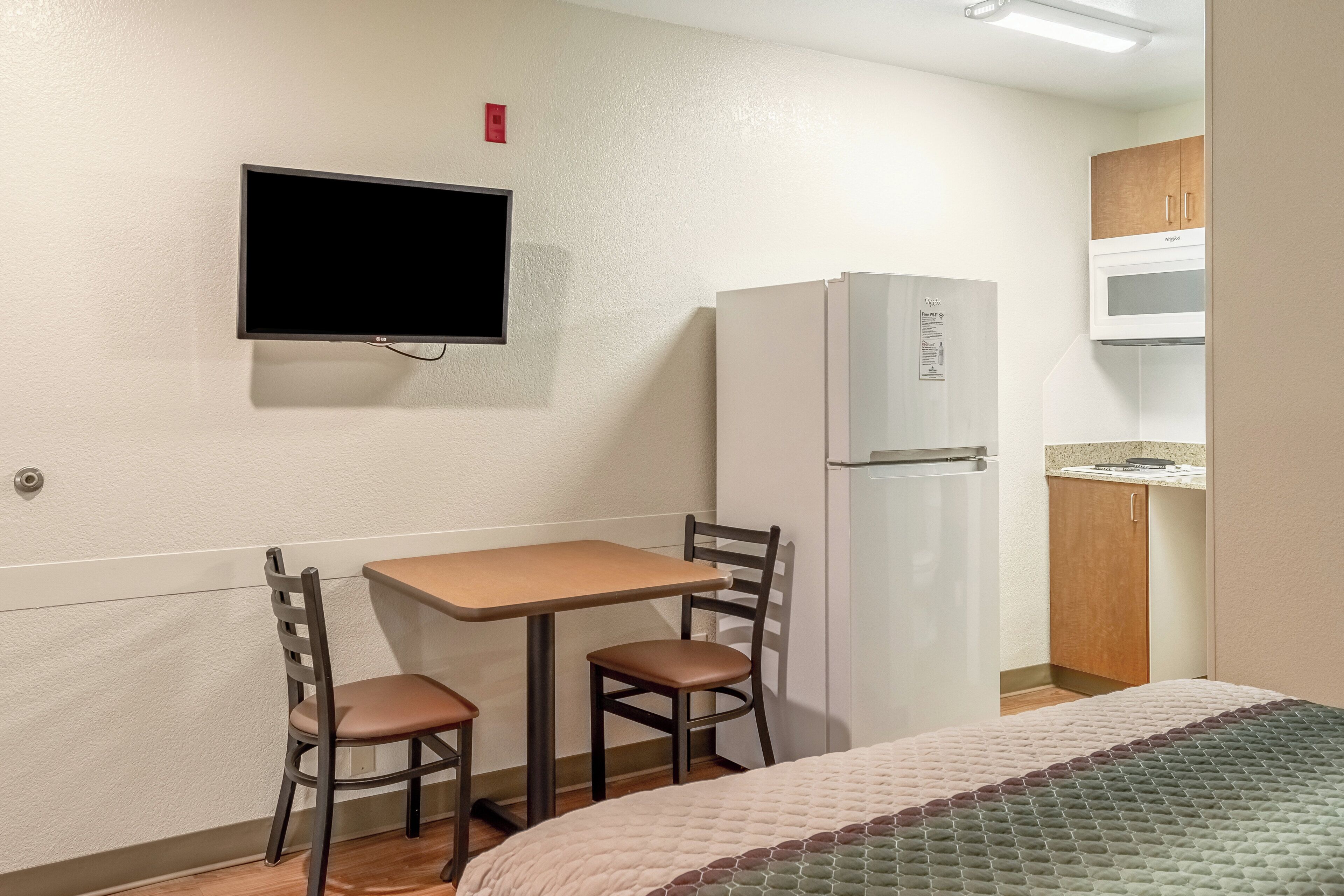 A small hotel room with a bed, a desk with two chairs, a television mounted on the wall, a refrigerator, and a microwave.