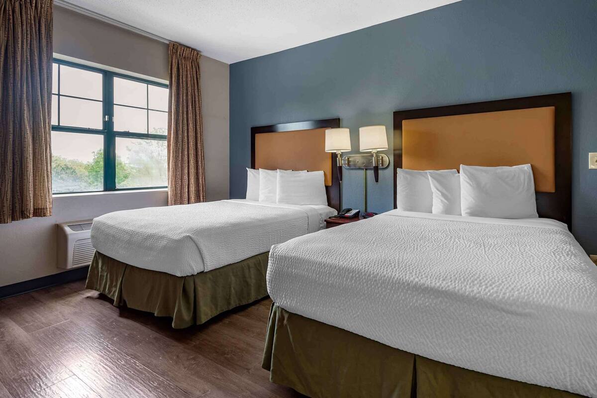 A hotel room with two beds, a window with curtains, a wall-mounted air conditioner, and a nightstand with a lamp.